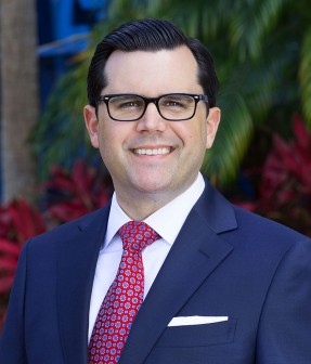 Portrait of a smiling man in a navy suit and standing outdoors with a building and plants in the background